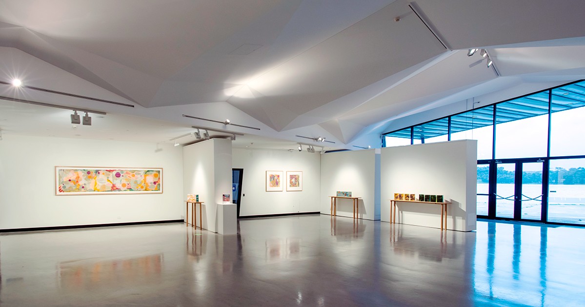 A photo of the Generator Gallery, a large open-plan space with art displayed on walls and stands, gallery lighting, and floor to ceiling windows that look out over Lake Ginninderra