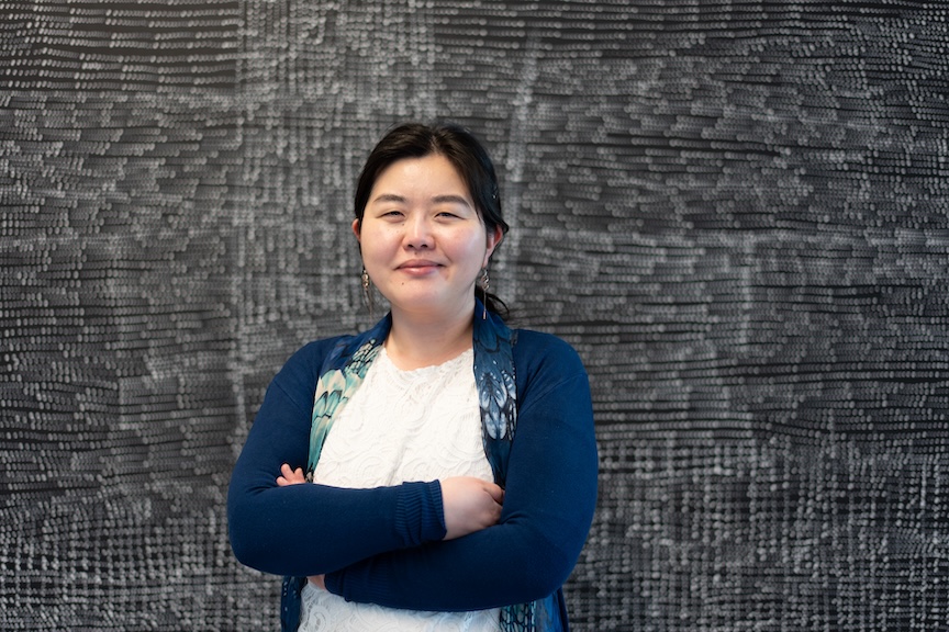 A photo of Emi Tanaka, standing in front of a large white-and-black artwork, arms folded and smiling at the camera