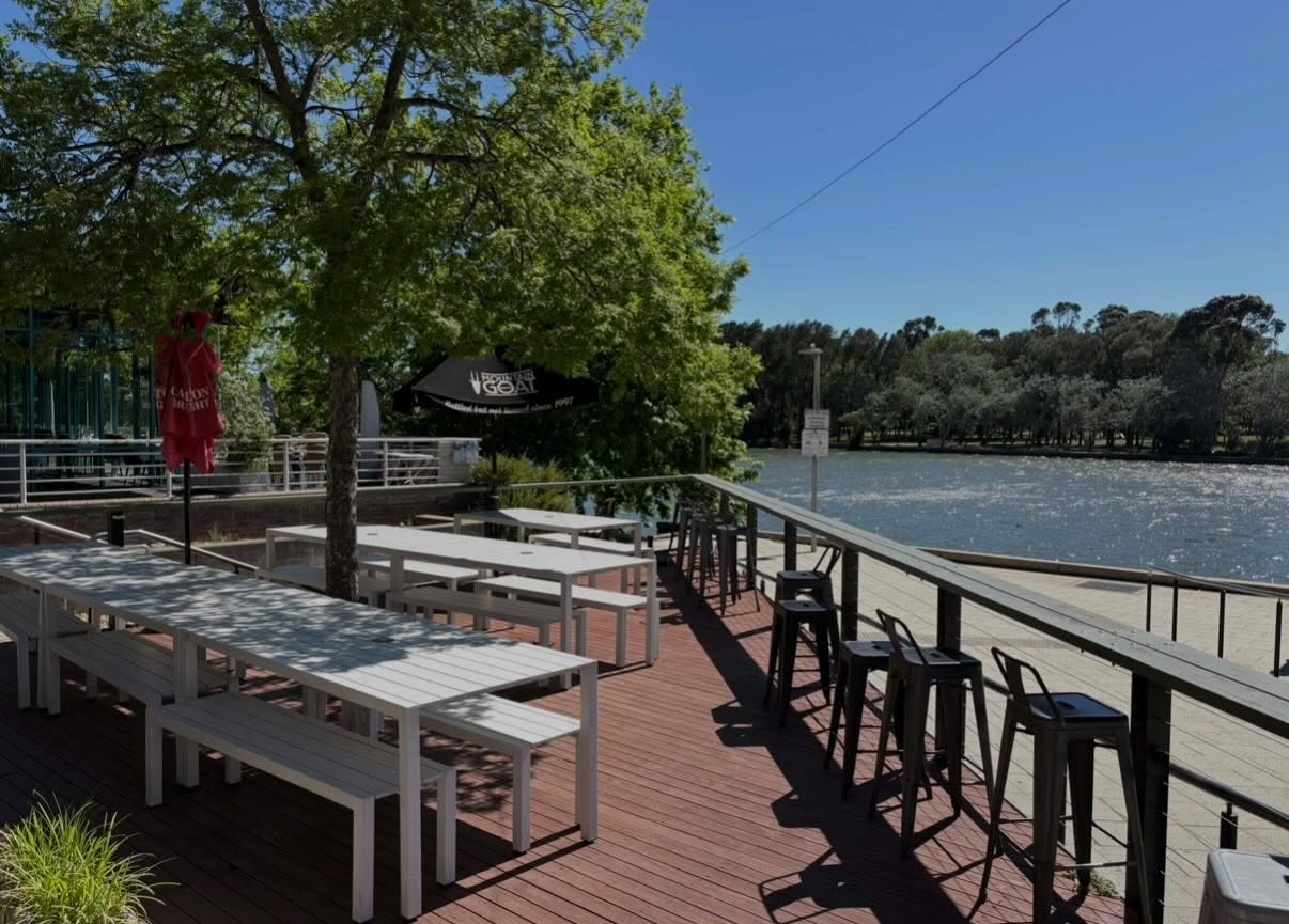 A photo of the Lighthouse Pub's beer garden. A decked area with seating shaded by trees, overlooking Lake Ginninderra